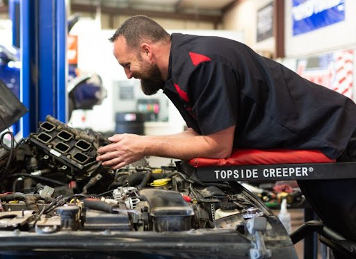 Mechanic working on car engine, leaning on a red topside creeper in a garage.