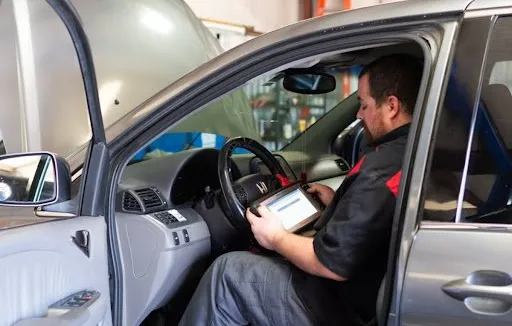 A technician in a uniform uses a diagnostic tablet inside a car with its hood open in a repair shop.