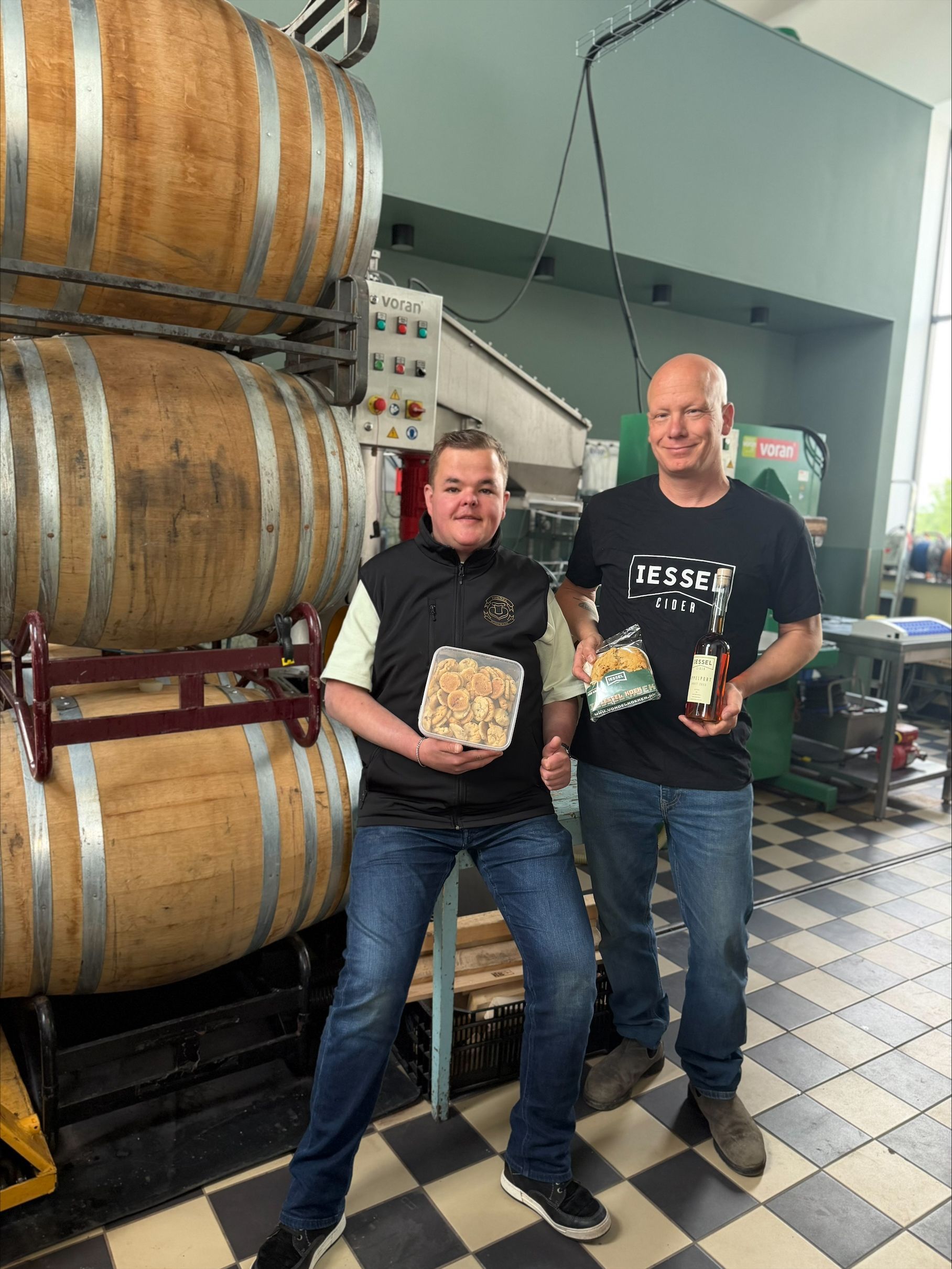 Twee mannen in de cidermakerij van IESSEL bij houten vaten, met een bak Vondelkoeken en een fles IESSEL appelport.