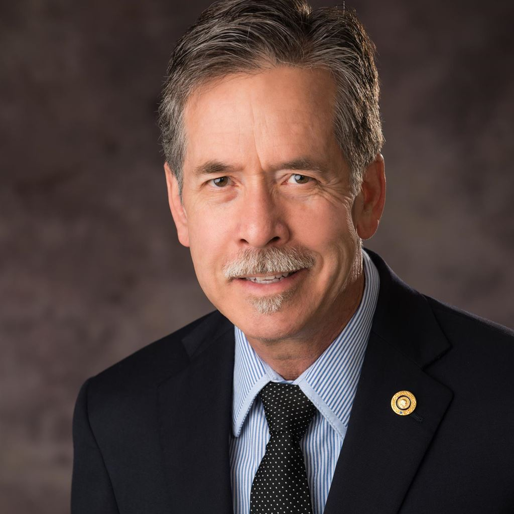 Headshot of a man in a dark suit and tie, smiling against a neutral studio background