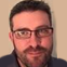 Close-up portrait of a bearded man wearing glasses, looking at the camera against a light background