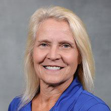 Portrait of a smiling person with long blonde hair wearing a blue shirt on a gray background