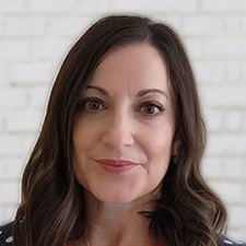 Woman with long brown hair in front of a white brick wall, looking at the camera with a slight smile