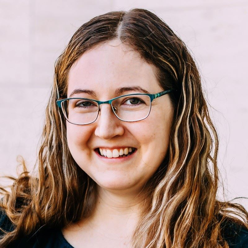 Smiling person with glasses and wavy brown hair against a light background