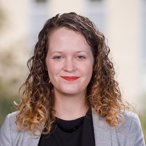 Smiling woman with curly hair in a gray blazer against a blurred indoor background