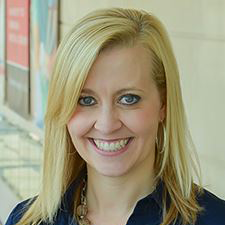 Smiling blonde woman in a navy blouse, standing indoors beside bookshelves.