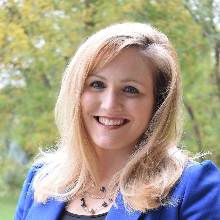 Smiling blonde woman in a blue blazer outdoors with blurred green foliage background