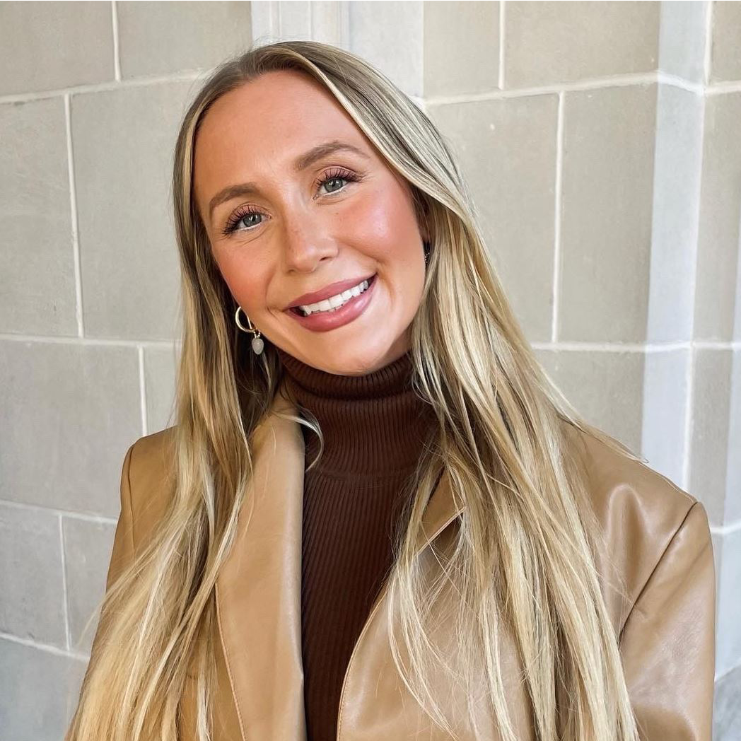 Smiling woman in a tan jacket and brown turtleneck standing against a light tiled wall