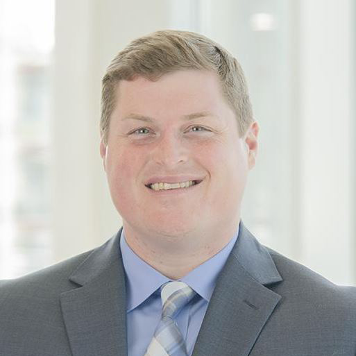 Smiling man in a gray suit and blue tie against a bright office background