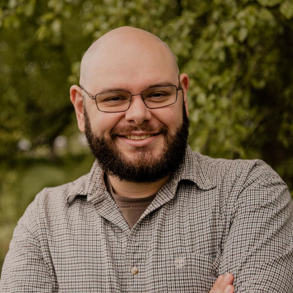 Smiling bald man with glasses and beard, arms crossed, outdoors against blurred greenery