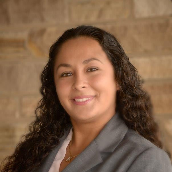 Smiling person in a gray blazer against a warm stone wall background