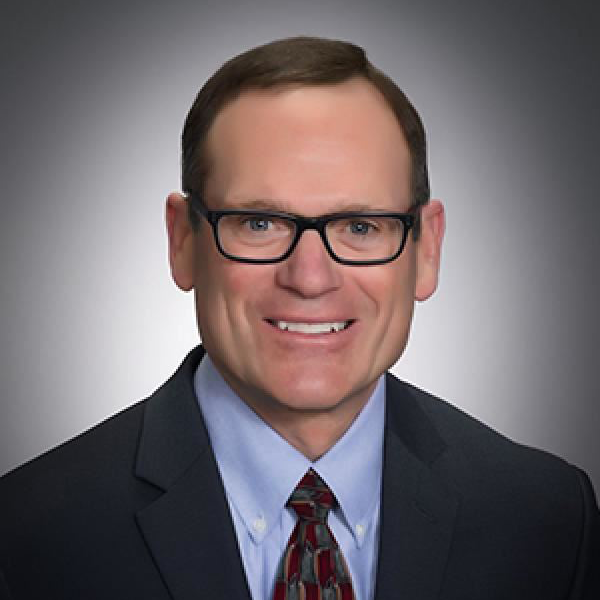 Professional headshot of a man in a suit, blue shirt, and glasses, smiling against a gray background