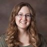 Smiling woman with long curly brown hair and glasses against a gray background