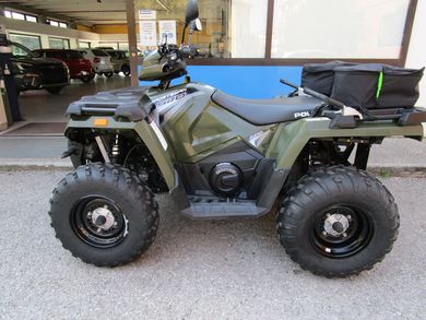 Un quad verde fuoristrada parcheggiato davanti a un negozio, con grandi pneumatici tassellati e una borsa portaoggetti posteriore.