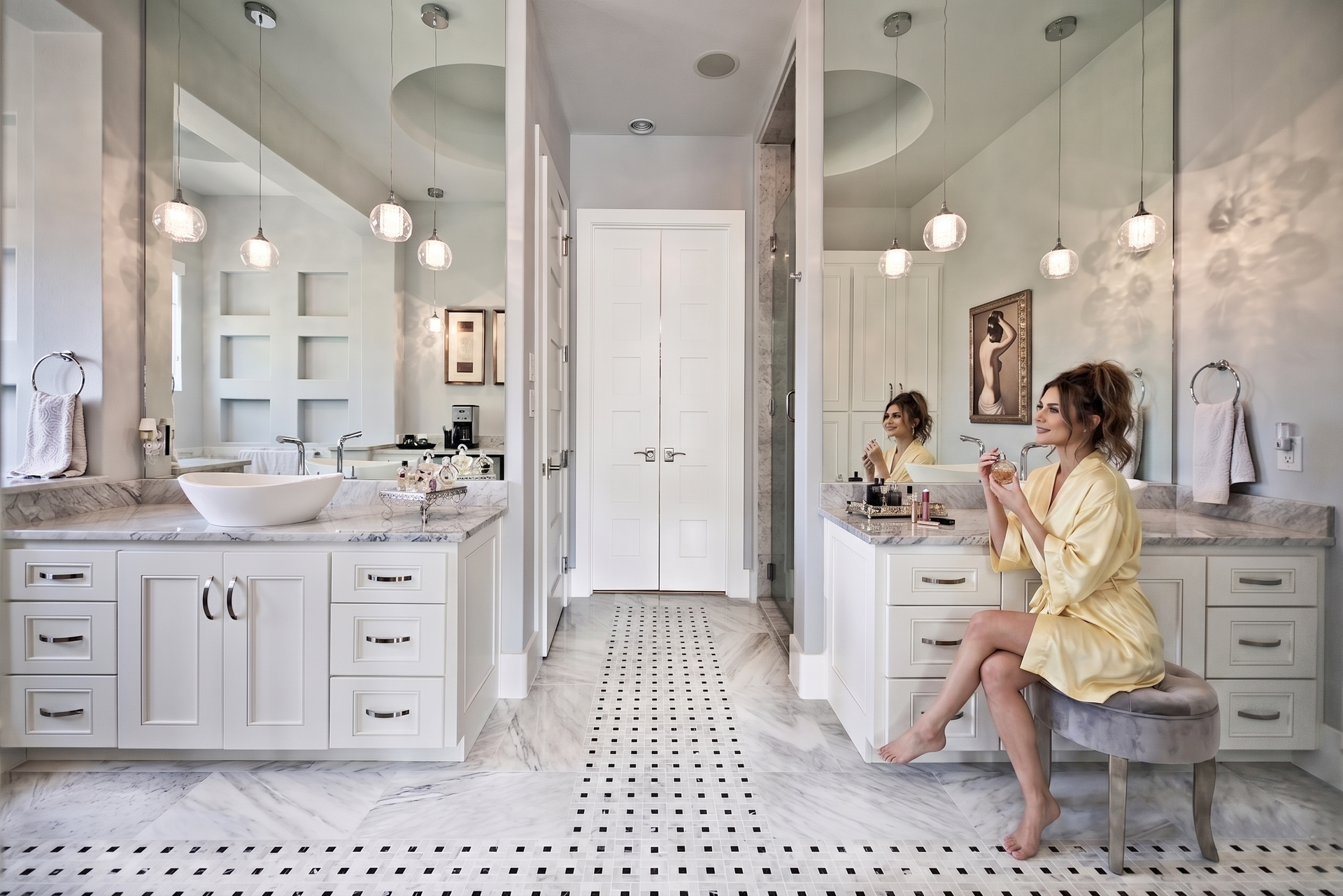 Bright white bathroom with double vanities and a women in a yellow robe sitting by Vanity mirror recently installed