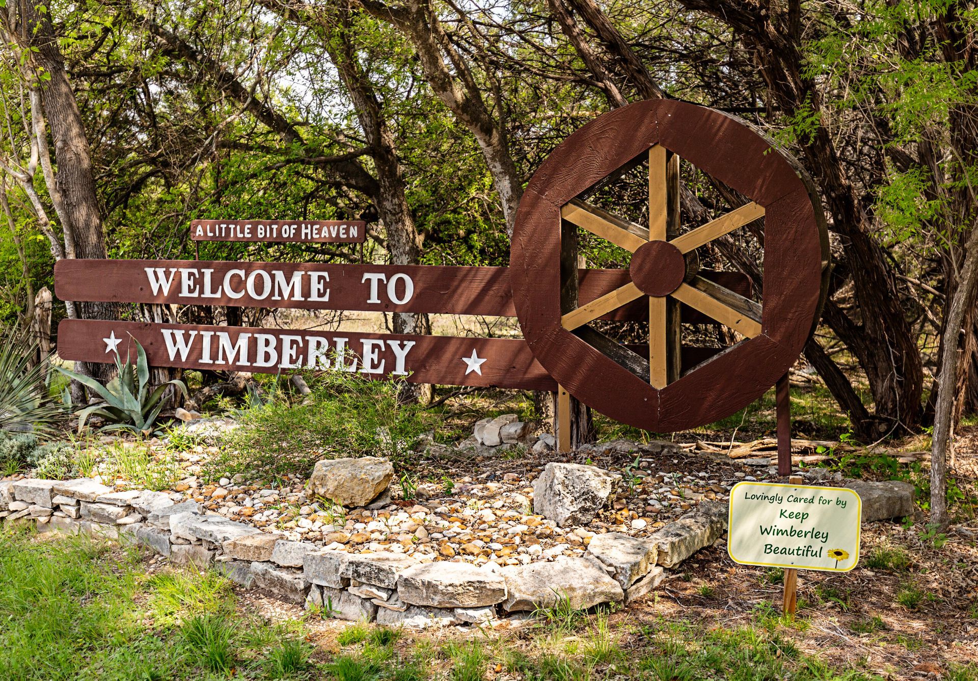 A welcome to wimberley sign with a wooden wheel in the background