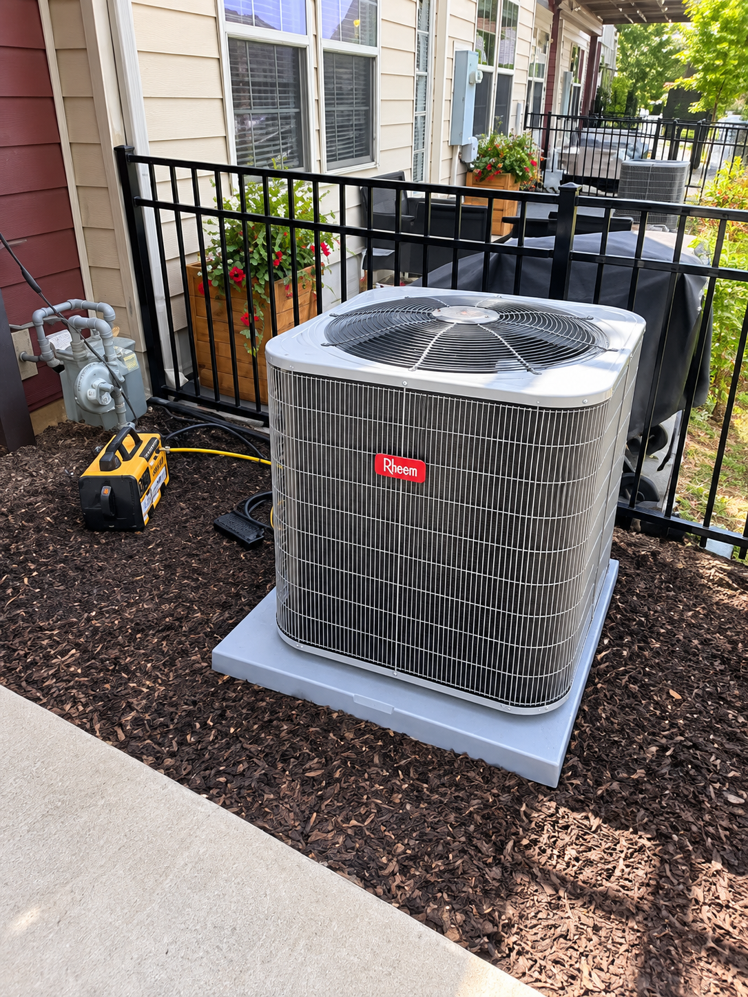 Outdoor air conditioner unit on a concrete pad beside a townhouse walkway, with a small tool nearby.