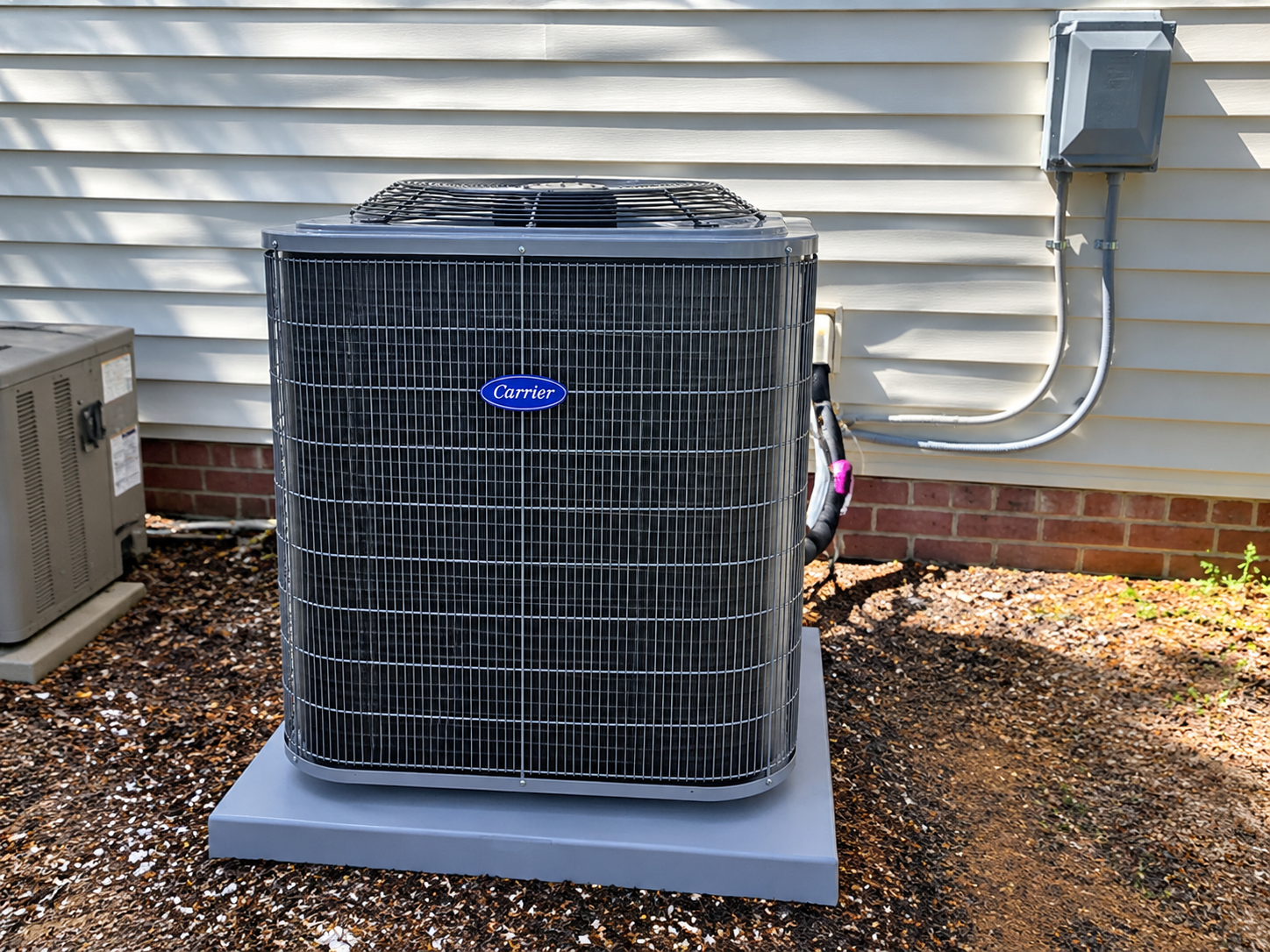 Outdoor air conditioner unit beside a house, with a black grille and gray pad.