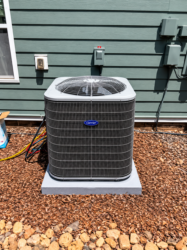 Outdoor air conditioner condenser on a gray pad beside a green house wall with mulch and rocks