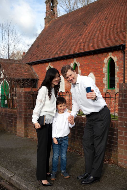 Family taking a selfie outside a red brick church. Woman smiles, holding a book. Son in jeans, man holds phone.