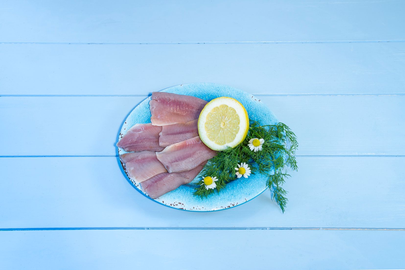 Ein blauer Teller mit Fischscheiben, Zitrone, Petersilie und Gänseblümchen auf einem blauen Holztisch.