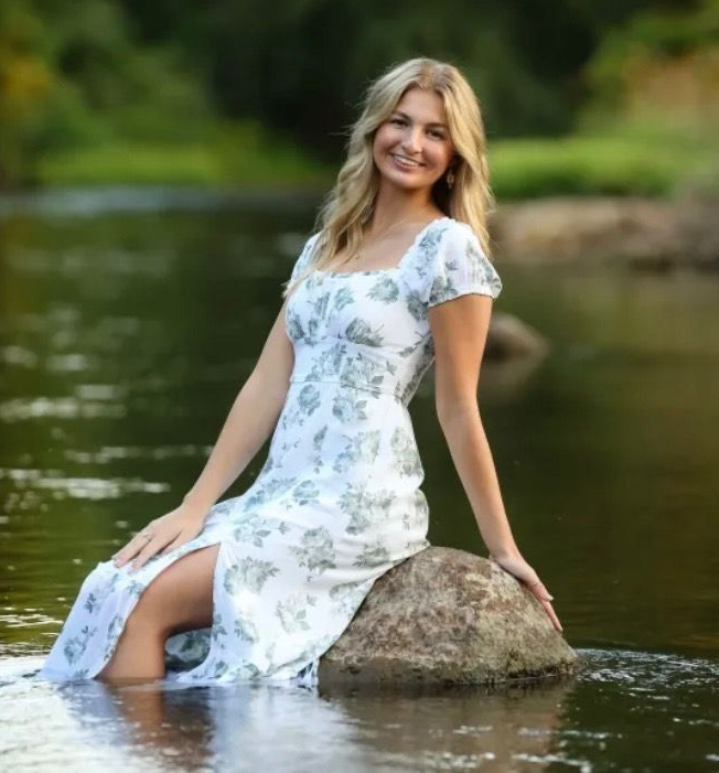 Blonde woman in white floral dress, sitting on a rock in a river, smiling at the camera.