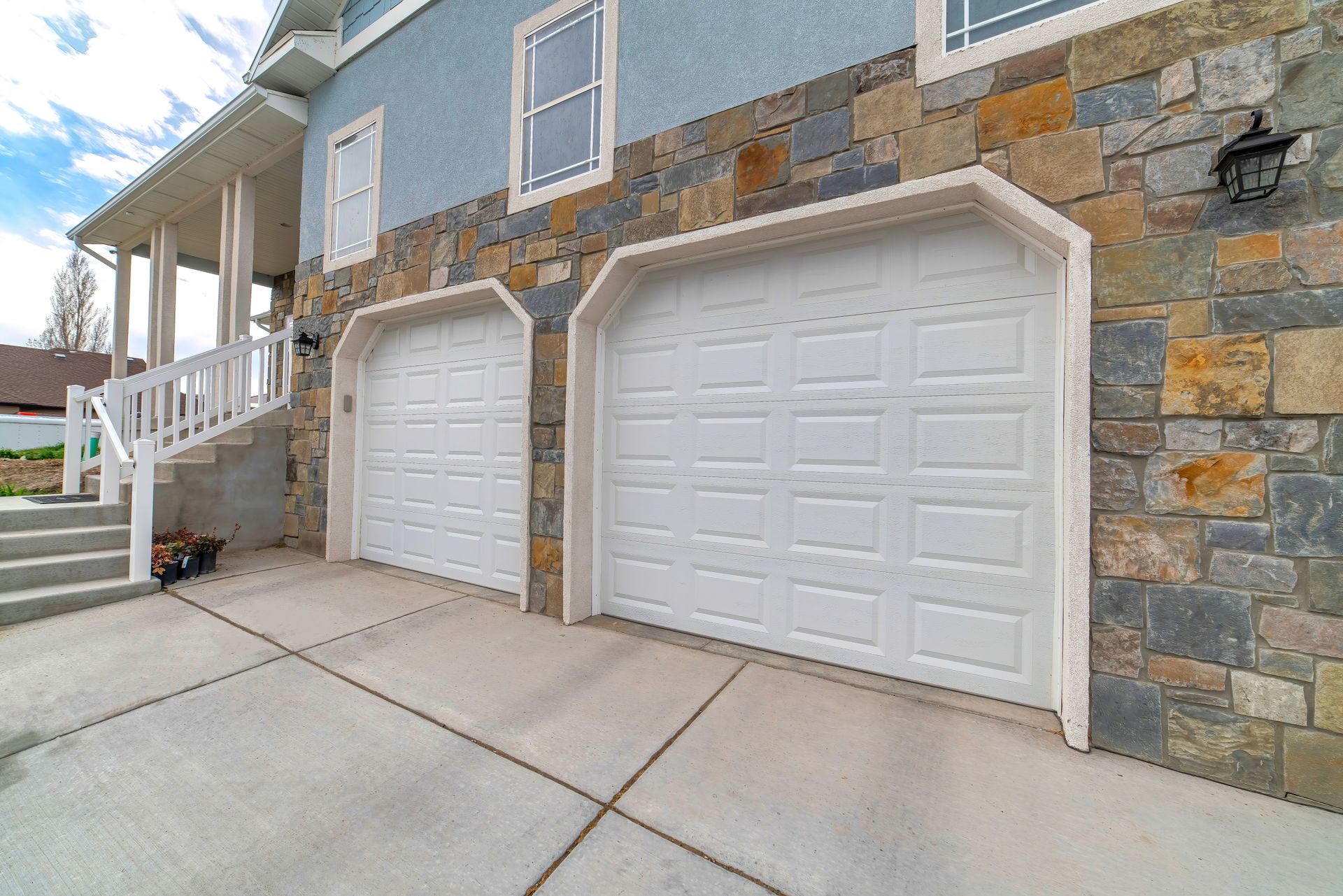 Two white garage doors on a stone-and-blue house with a concrete driveway and front steps