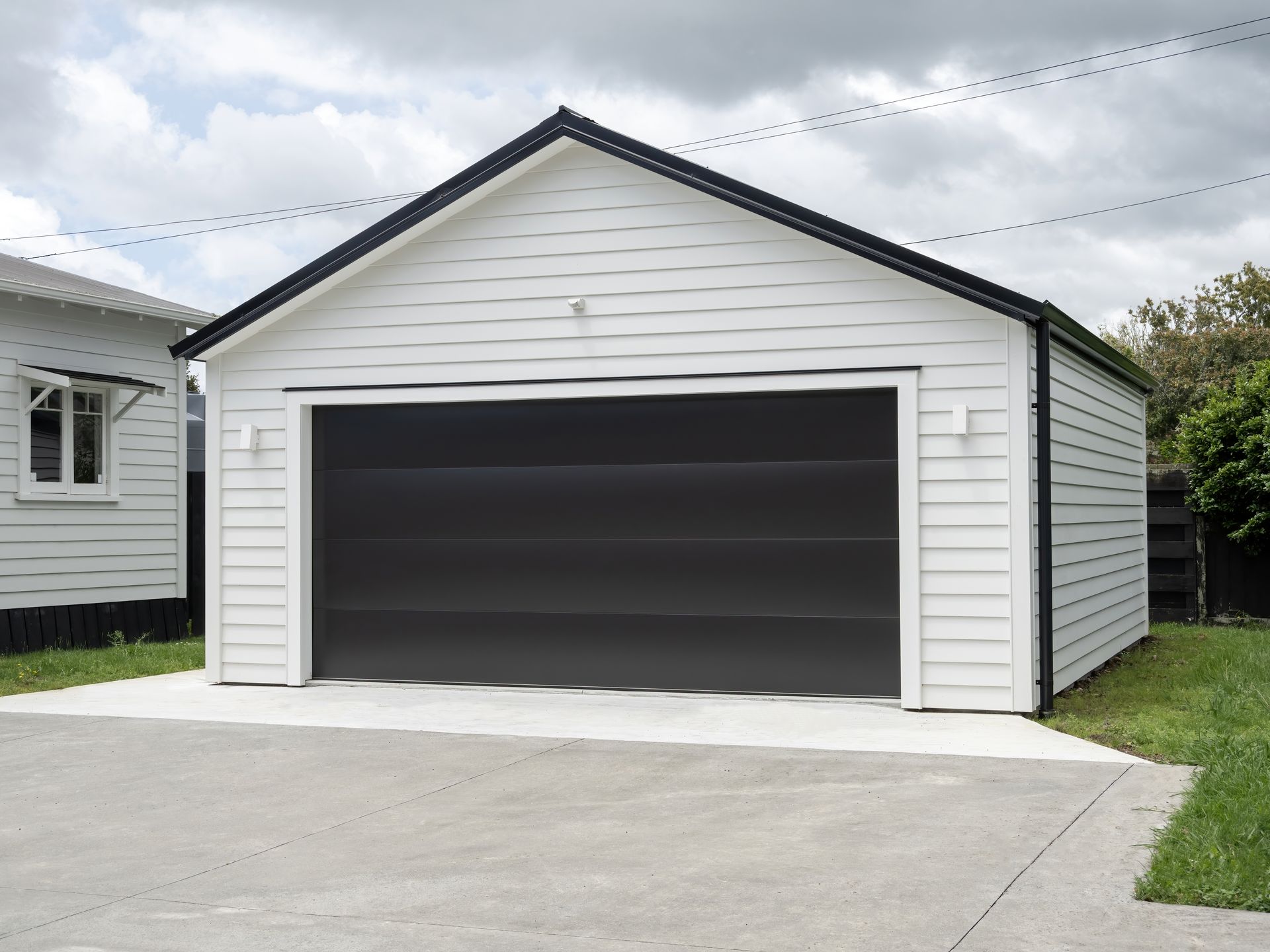 White detached garage with a large black door and concrete driveway under a cloudy sky
