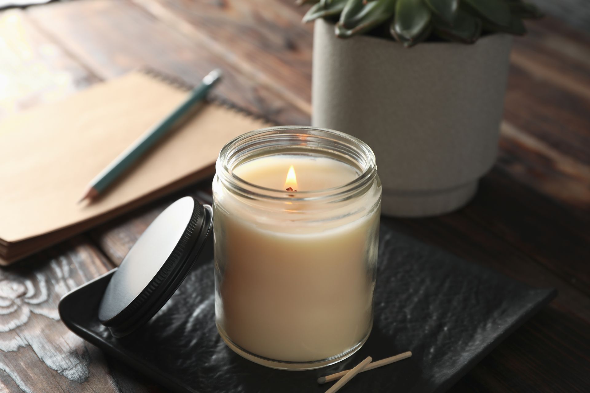 Lit beige candle in a glass jar on a black plate beside a spoon and notebook on a wooden table