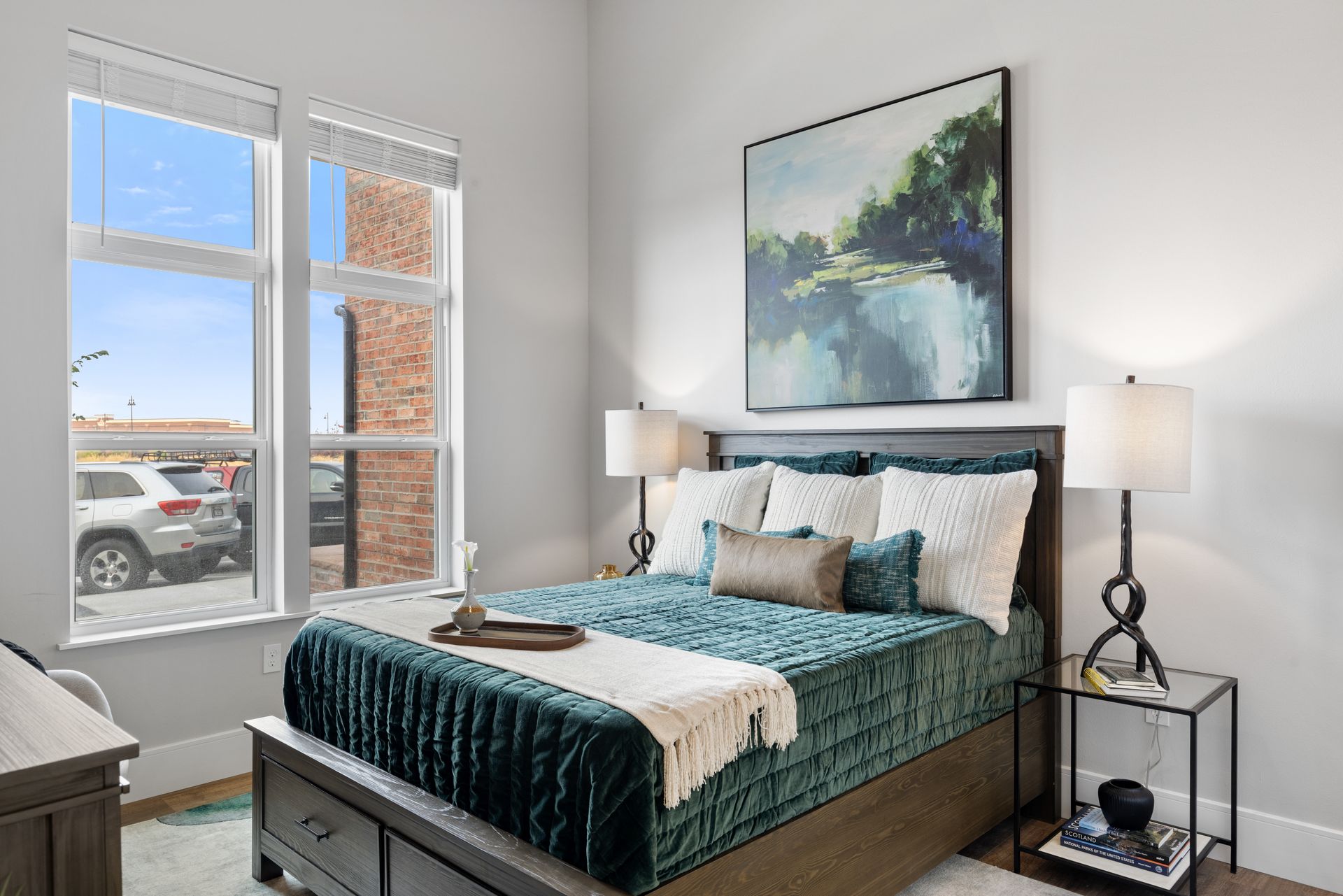 Bright bedroom with a made bed, teal bedding, artwork, lamps, and large windows.