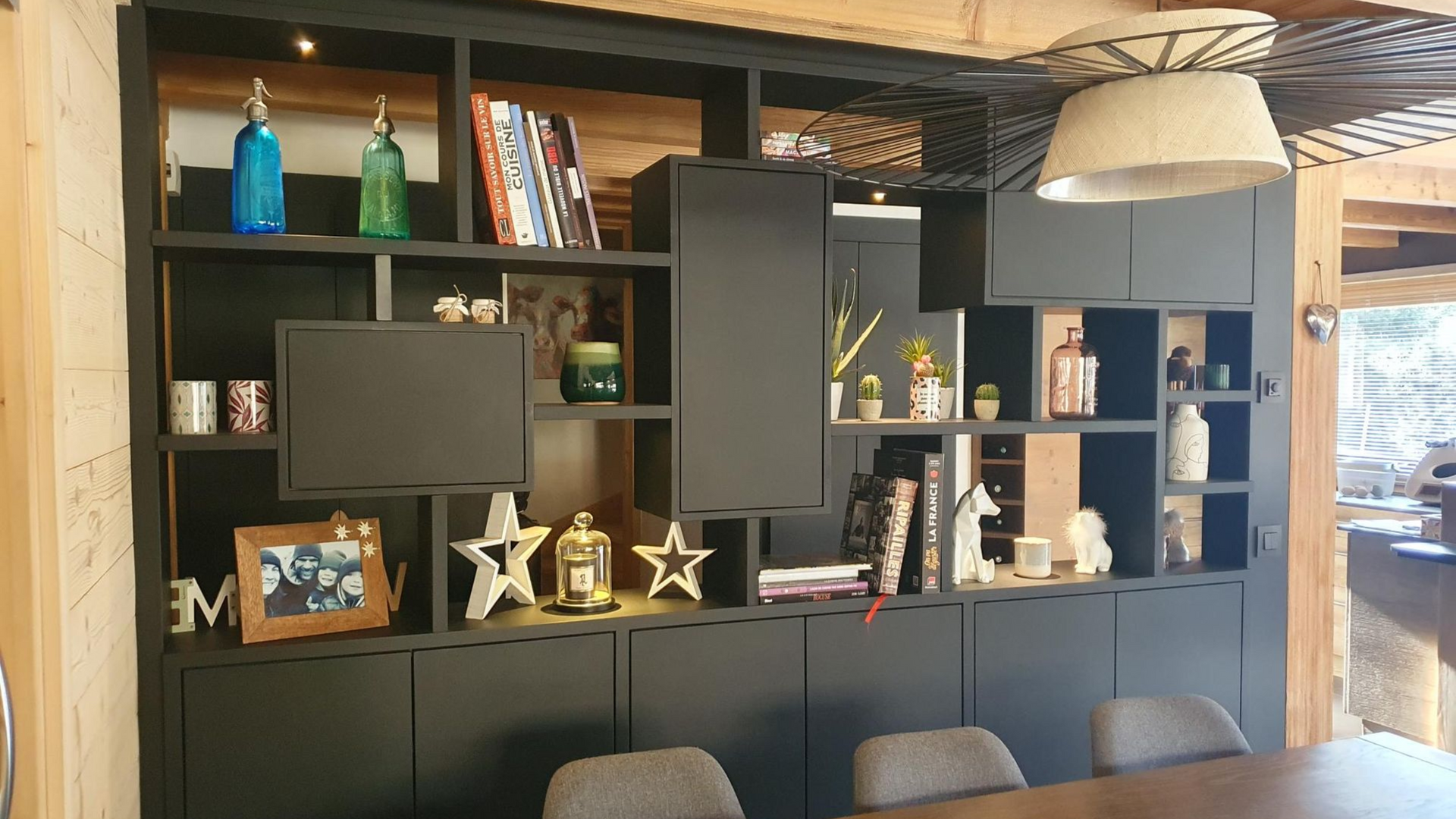 Meuble encastré noir avec étagères ouvertes et placards fermés, présentant livres et objets de décoration. Table et chaises marron devant.