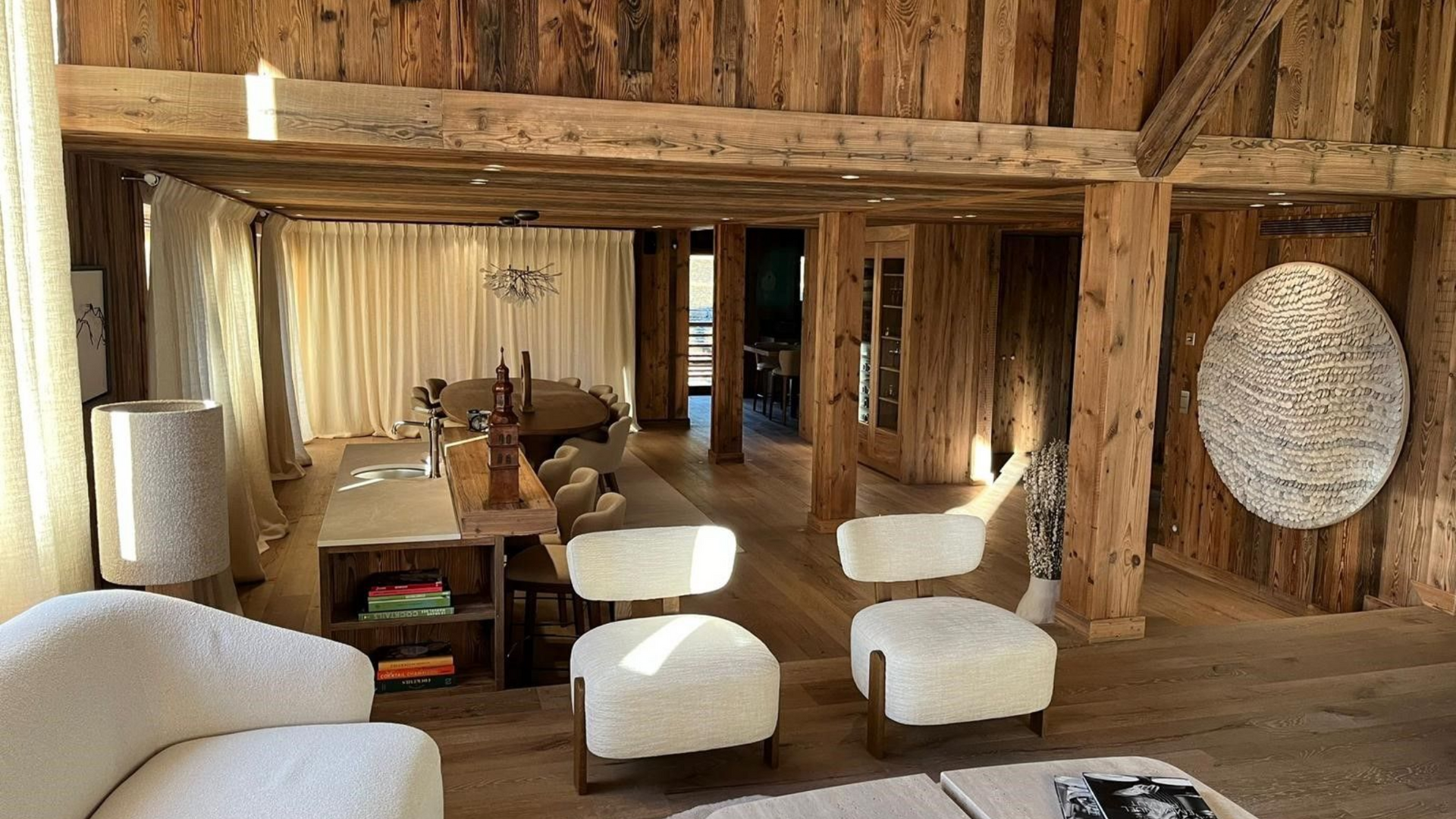 Intérieur chaleureux avec murs et meubles en bois, comprenant des chaises blanches et une grande tenture murale texturée.