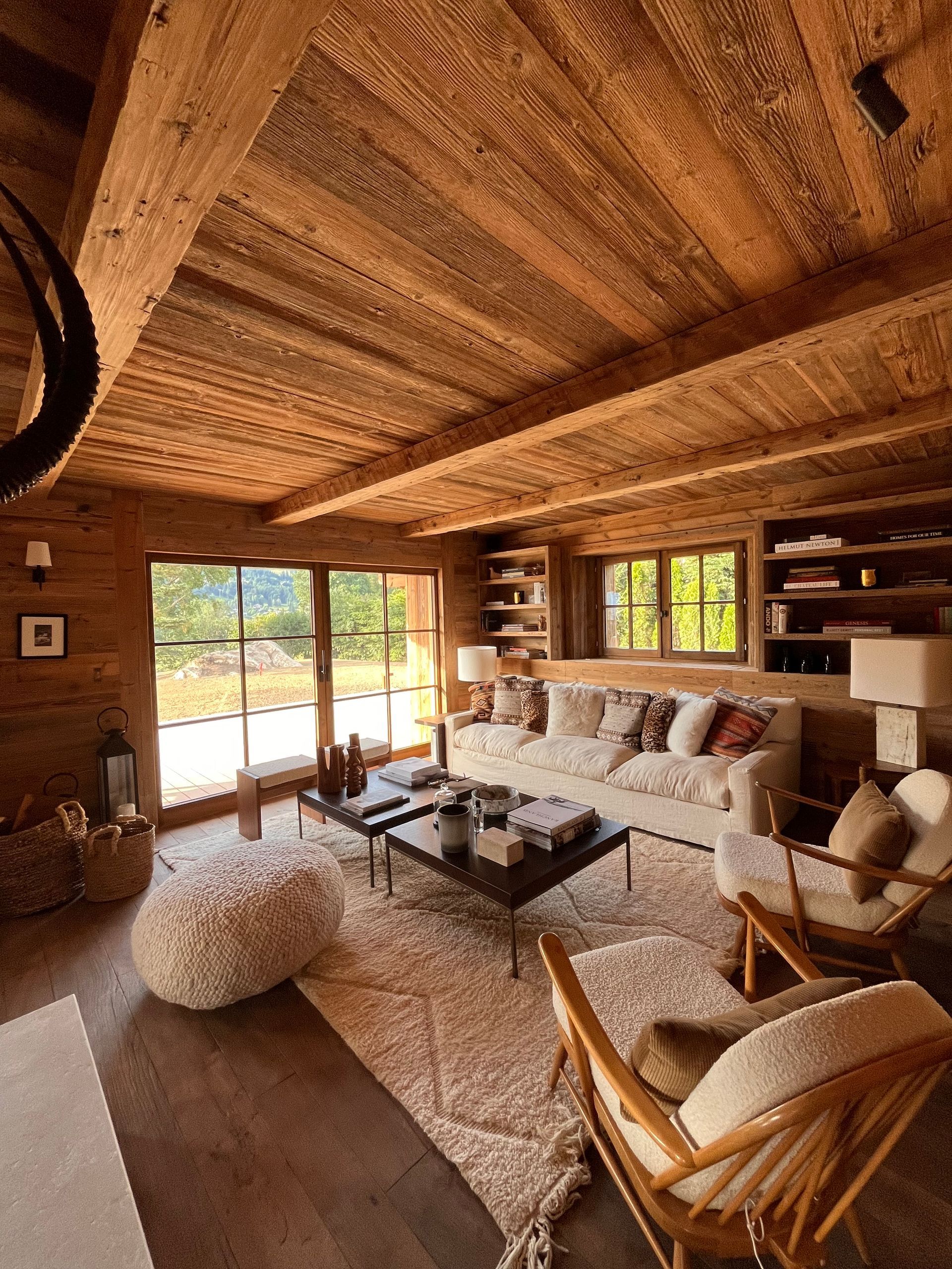 Salon chaleureux avec plafond et murs en bois. Canapé, fauteuils et tapis beiges, avec vue sur l'extérieur.