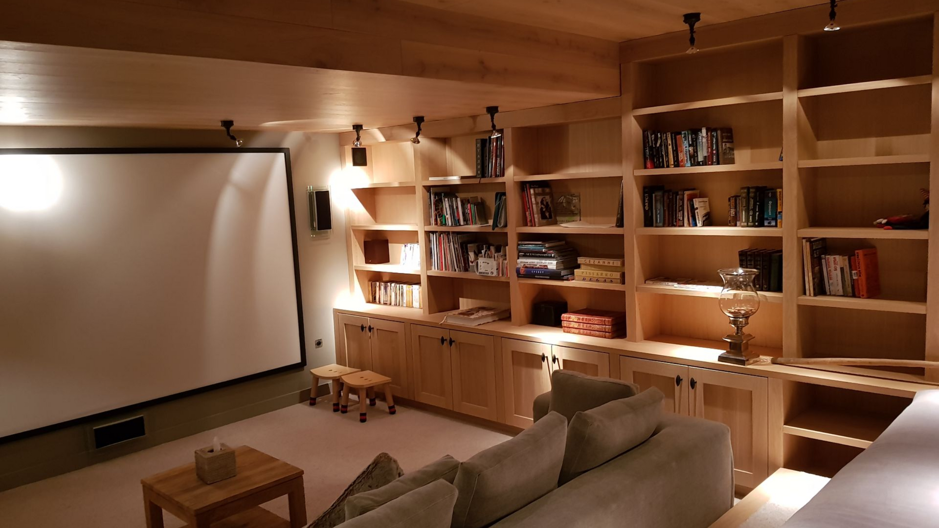 Salle de cinéma maison avec étagères en bois intégrées remplies de livres et de supports multimédias. Un écran de projection est accroché à gauche.