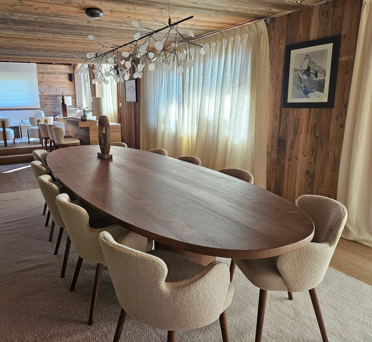 Table de salle à manger ovale en bois avec chaises dans une pièce rustique aux murs en bois et aux rideaux drapés.