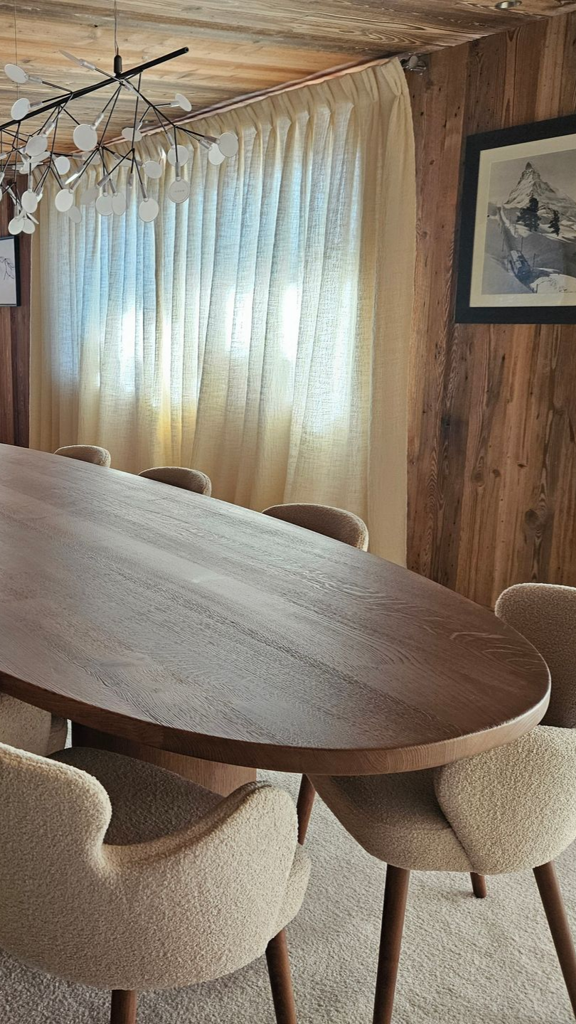 Table de salle à manger ovale en bois avec chaises dans une pièce lambrissée, fenêtre avec voilages.