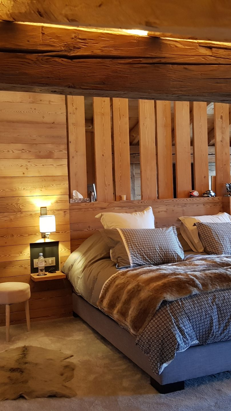 Chambre avec murs en bois, lit, couverture en fourrure, coussins décoratifs et table de chevet intégrée avec lampe.