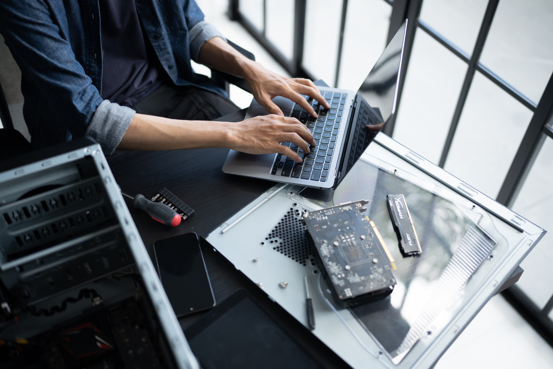 Technician typing on a laptop beside a disassembled computer on a workbench.