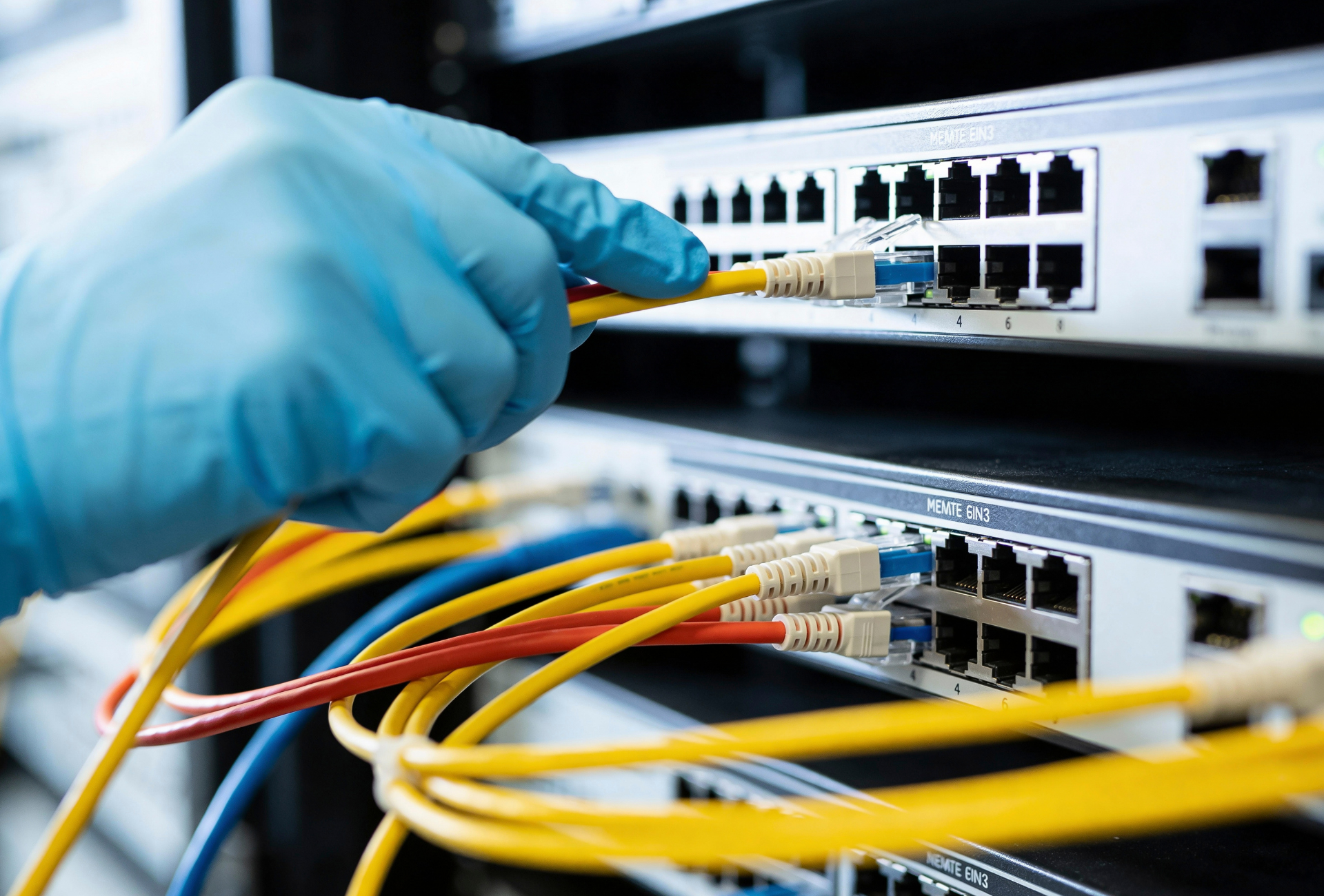 Gloved hand plugging a yellow Ethernet cable into a network switch with other cables attached