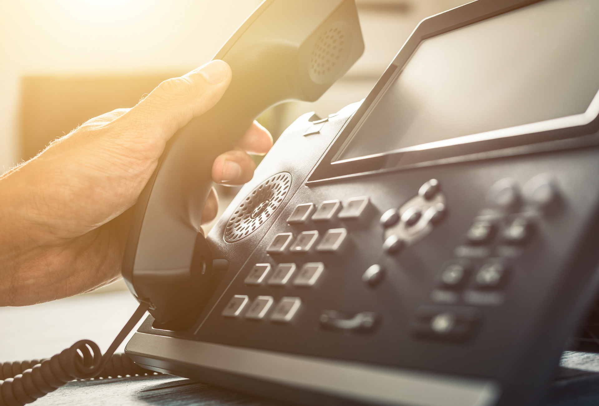 Hand holding office phone handset over desk phone keypad in warm light