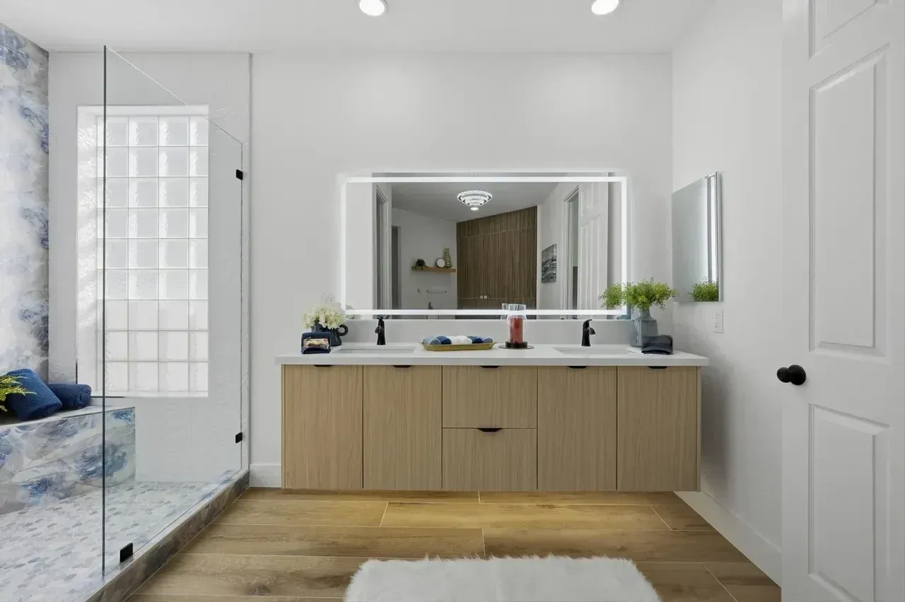 Modern bathroom with light wood vanity, illuminated mirror, shower, and white walls.