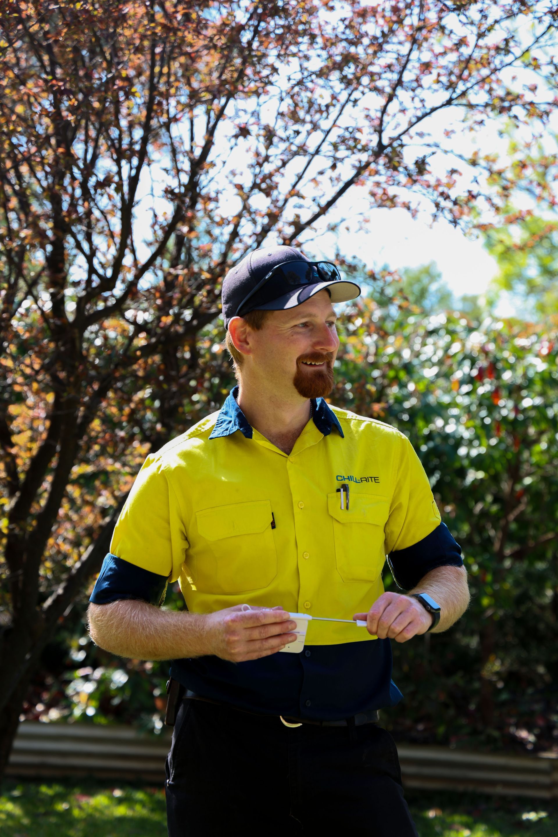 Man in yellow and navy work shirt and cap, holding something, looking away, outdoors— Chill-Rite Refrigeration and Air Conditioning in Dubbo, NSW