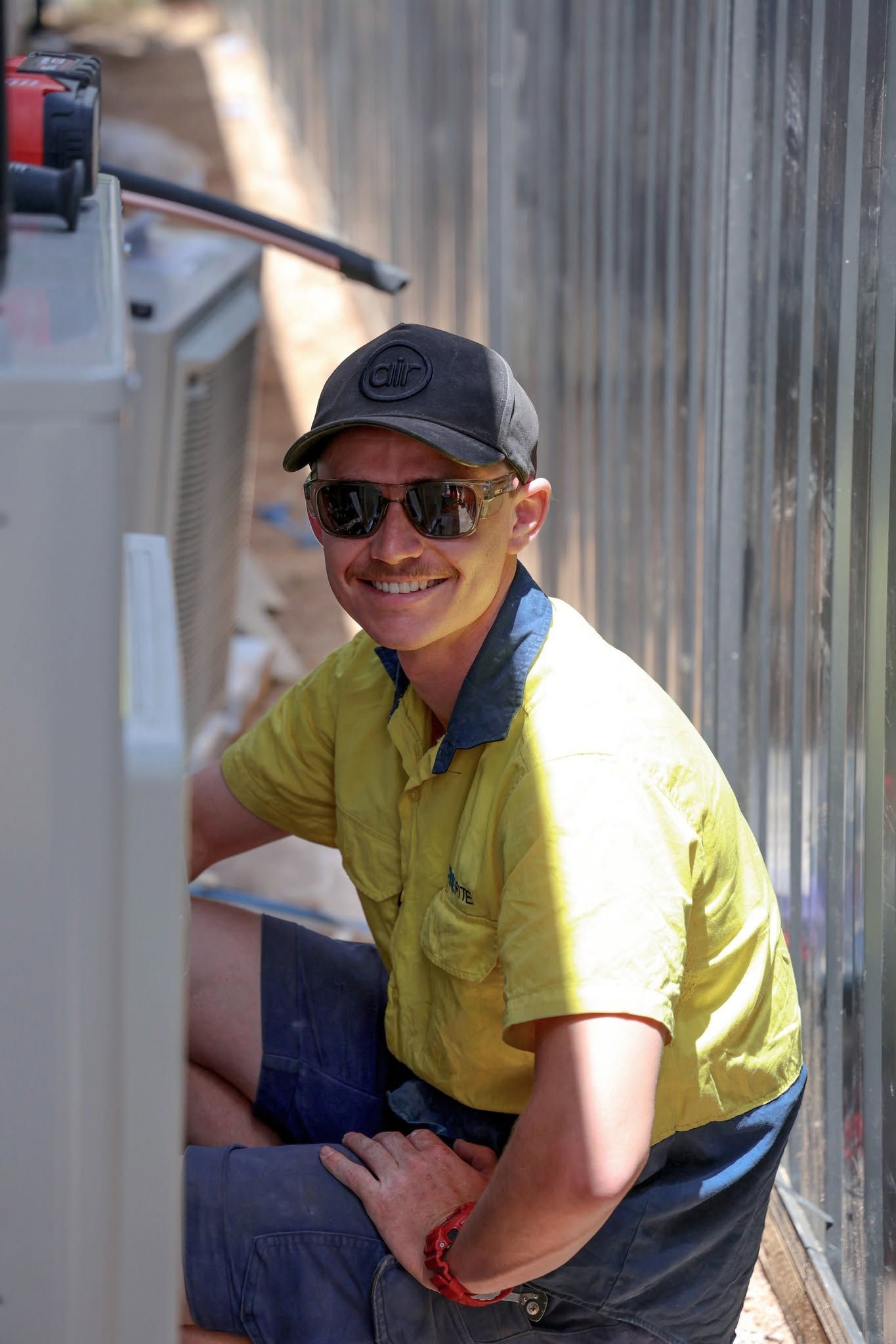 Smiling Worker in Yellow Shirt and Cap Crouches Beside a Metal Wall — Chill-Rite Refrigeration and Air Conditioning in Wodonga, VIC