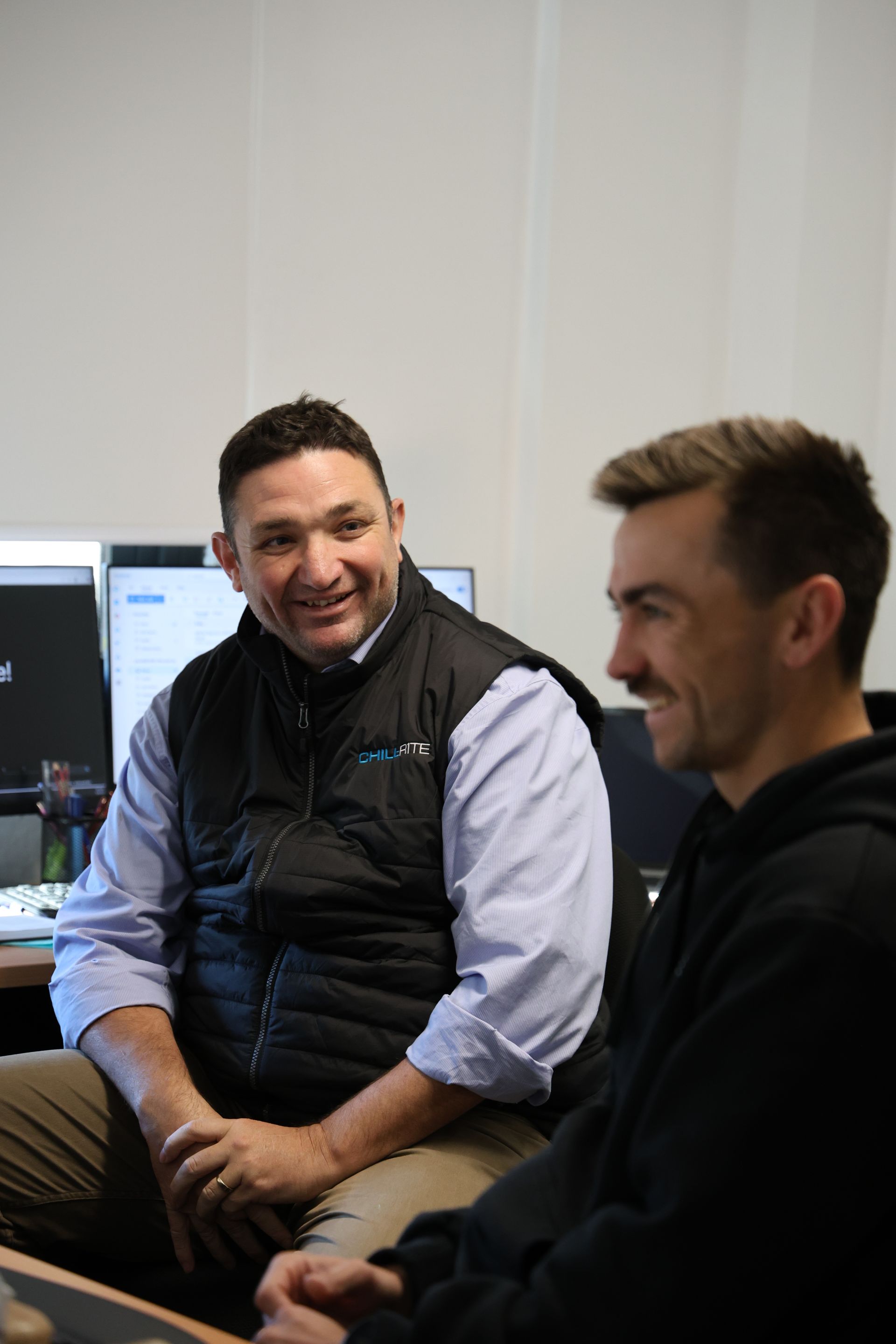 Two men smiling, in an office setting. One in a vest, the other in a hoodie, near computers— Chill-Rite Refrigeration and Air Conditioning in Newcastle, NSW