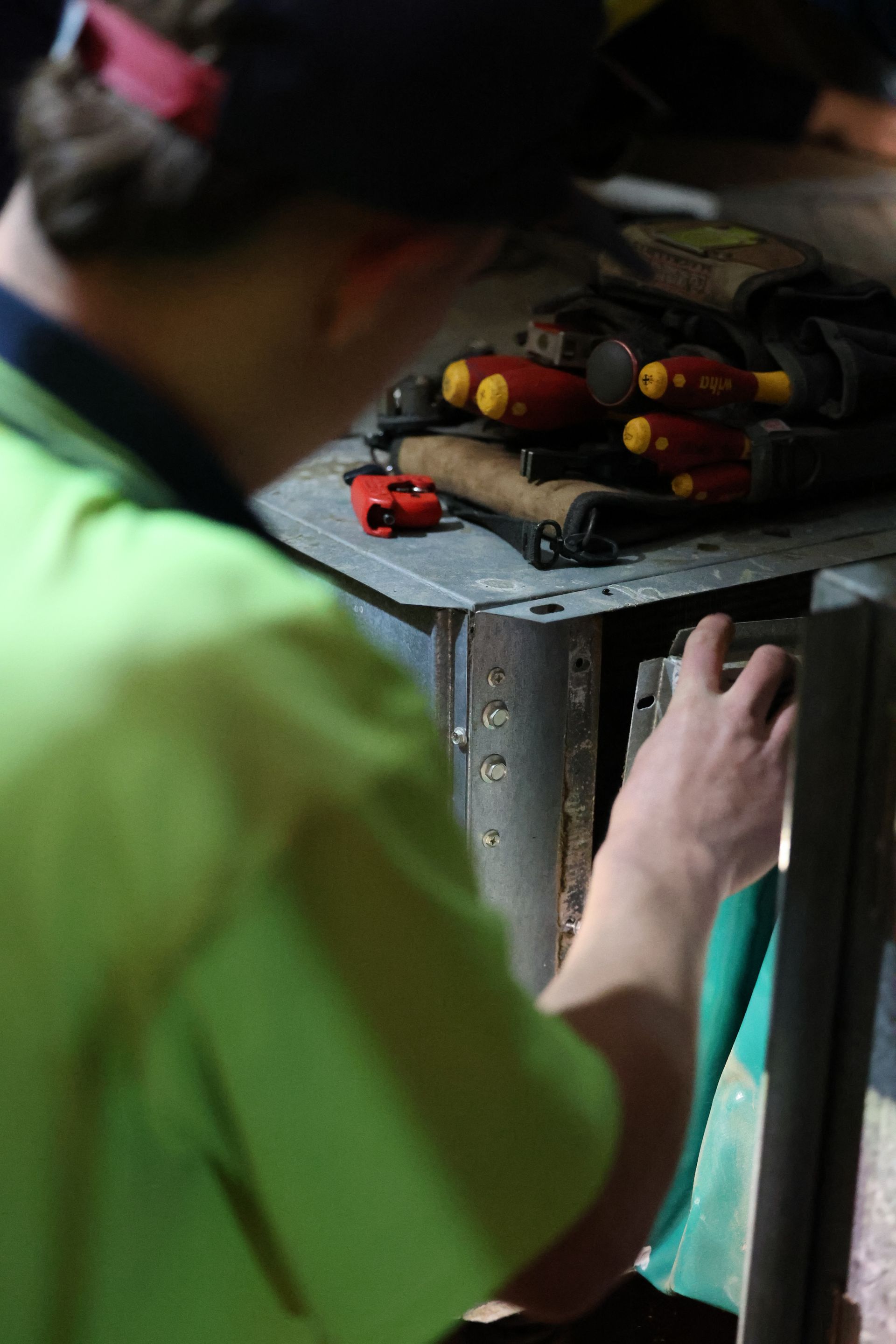 Electrician in a green shirt working on a metal panel, tools in pouch visible— Chill-Rite Refrigeration and Air Conditioning in Newcastle, NSW