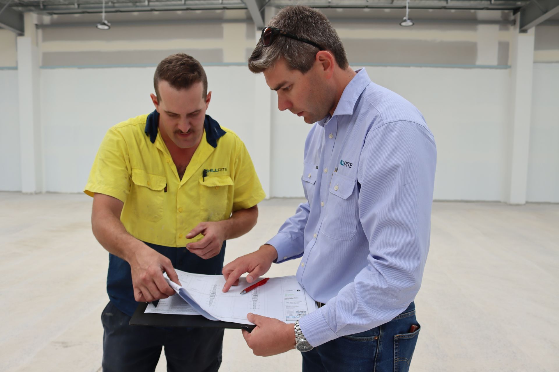 Two men reviewing blueprints in an empty industrial building, one pointing, wearing work clothes— Chill-Rite Refrigeration and Air Conditioning in Coffs Harbour, NSW