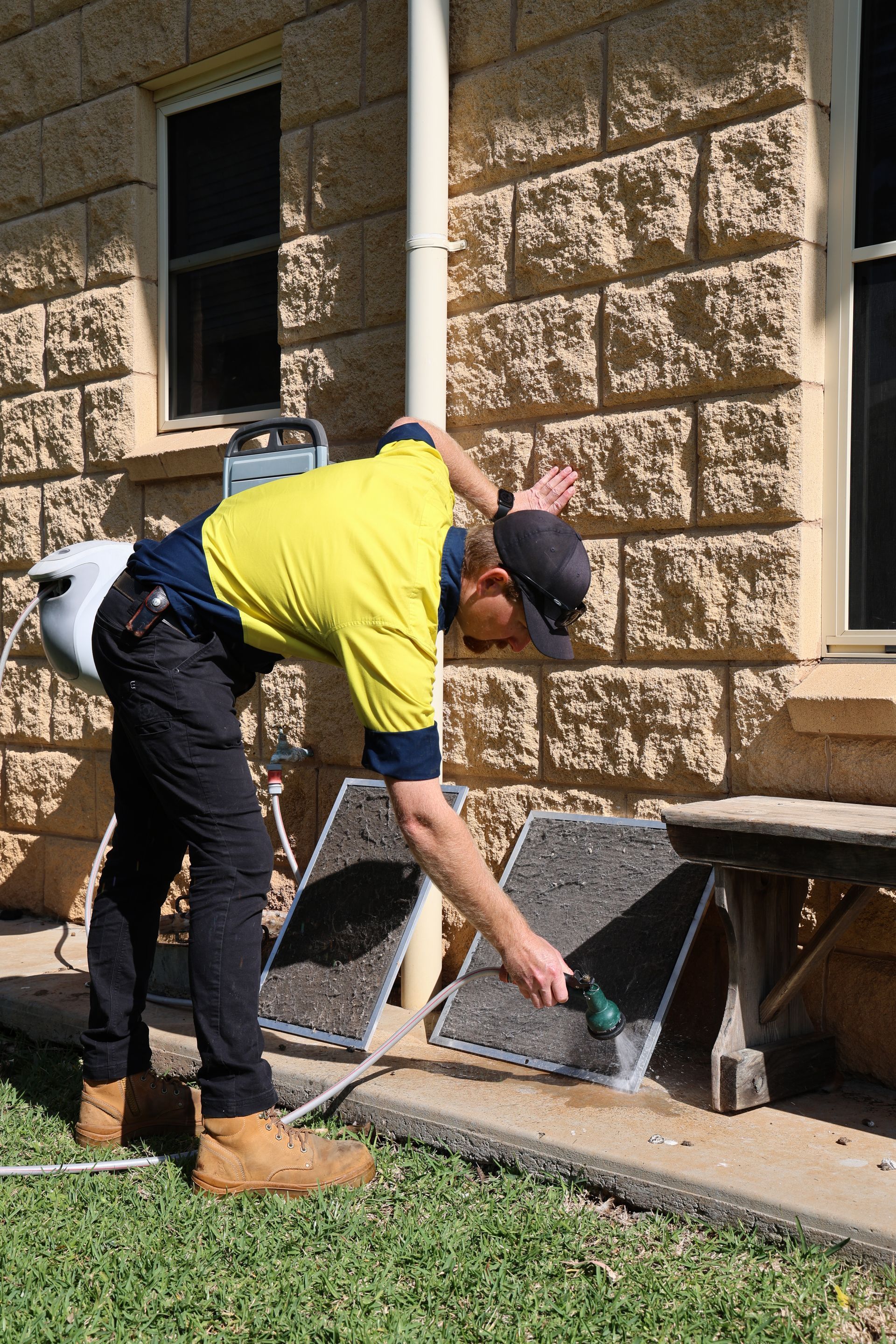 Man washing two dark grilles with a hose outside a brick building— Chill-Rite Refrigeration and Air Conditioning in Dubbo, NSW