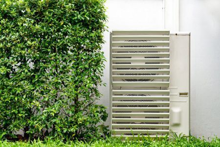 A White Air Conditioner is Sitting on the Side of a Building Next to a Bush — Chill-Rite Refrigeration and Air Conditioning in Dubbo, NSW