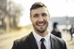 Man in a black suit smiles broadly outdoors on a sunny day.