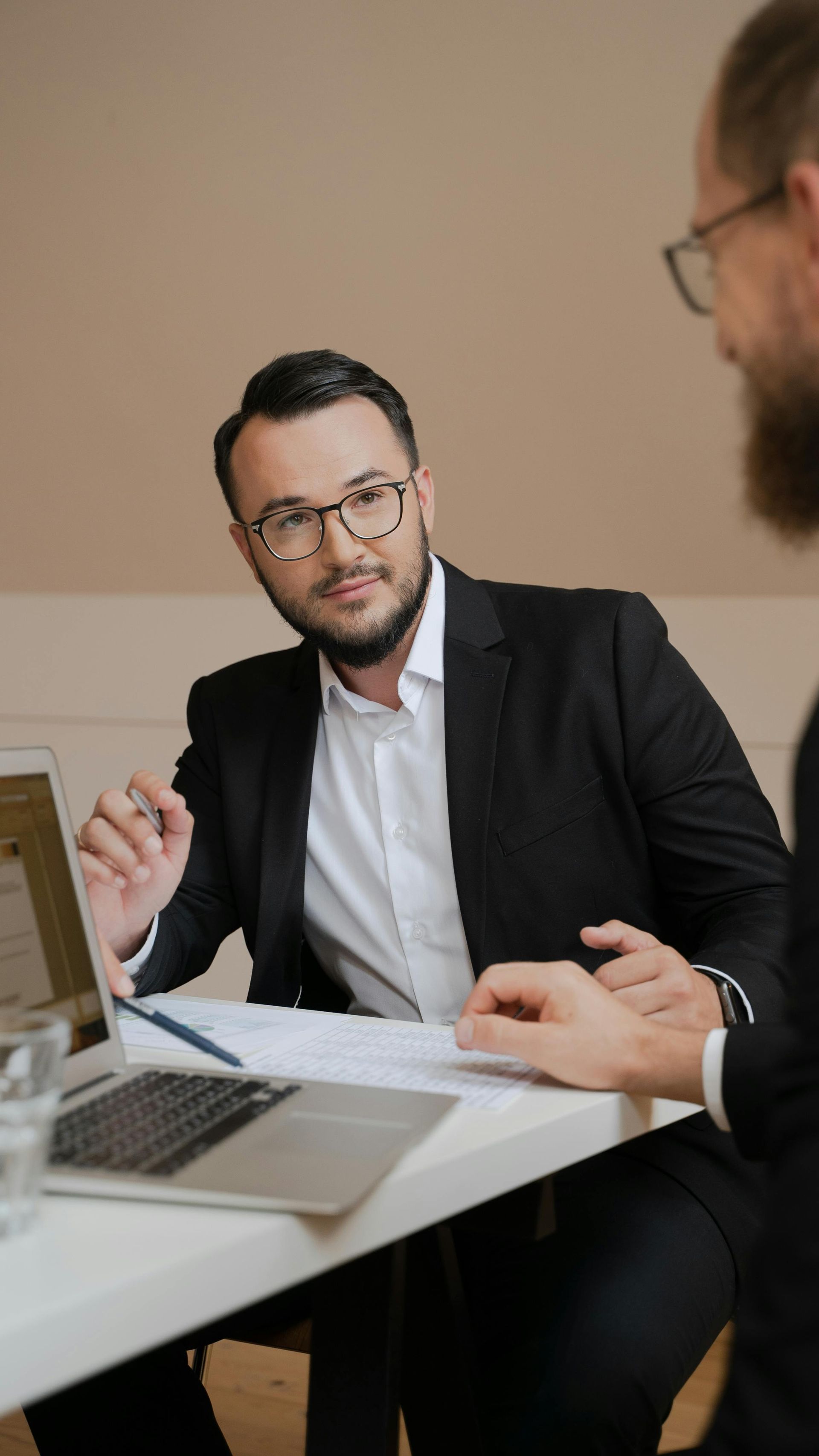 Two men in suits at a table with papers and a laptop, one gesturing and looking at the other.